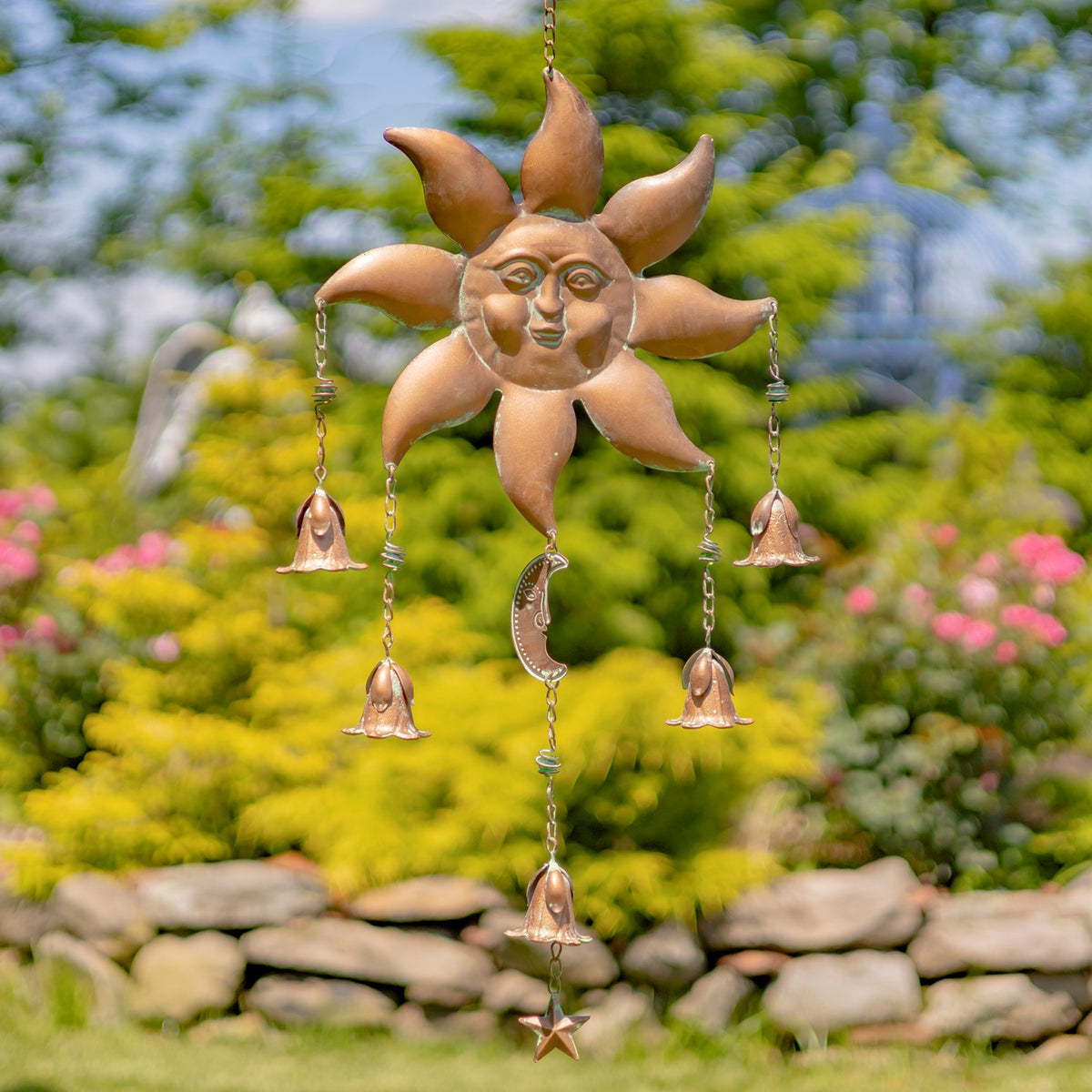 A sun-faced wind chime with curved rays hangs outdoors, with chains, bells, a small moon and a star.