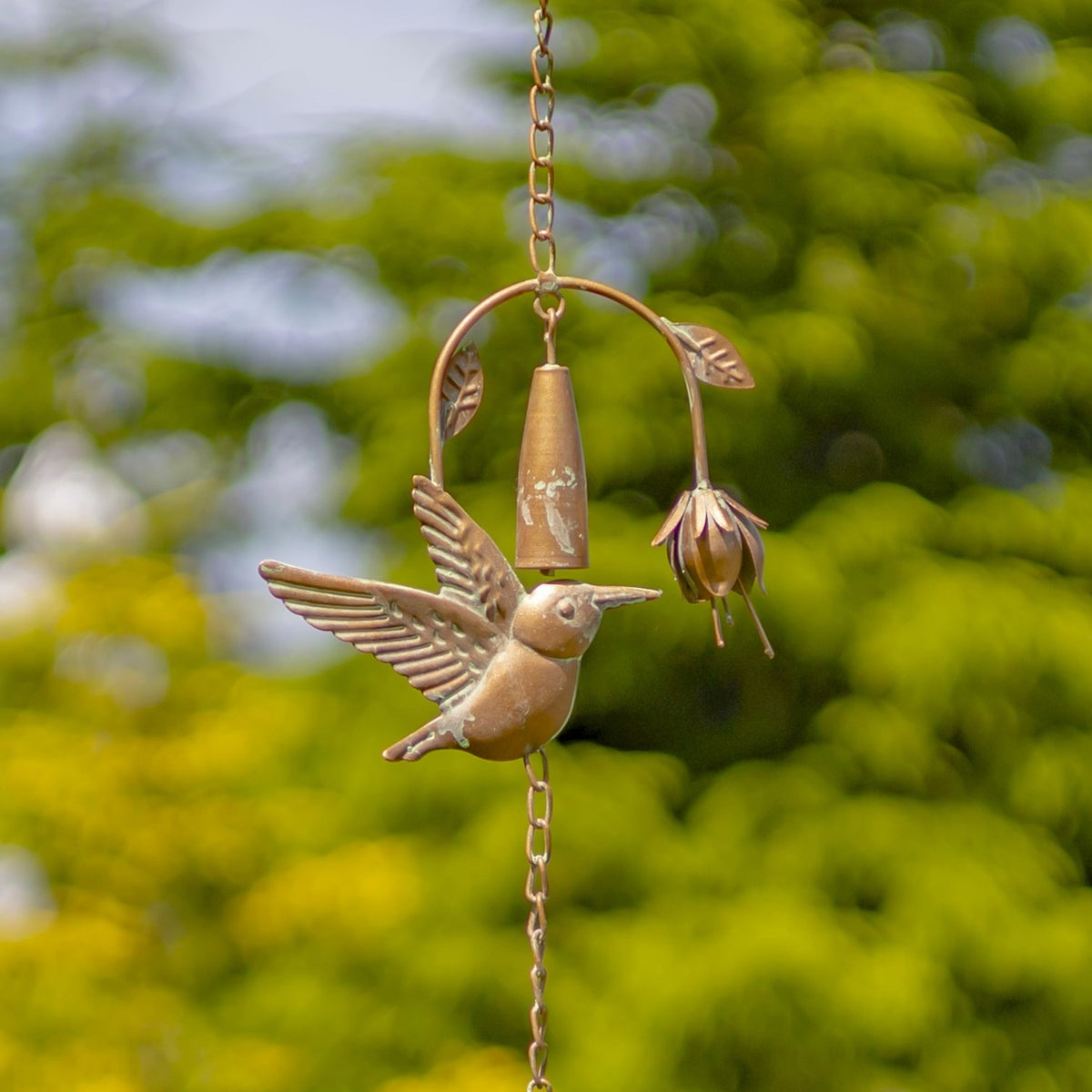 Close view of a metal hummingbird with raised wings beside a flower-shaped bell on the chain.