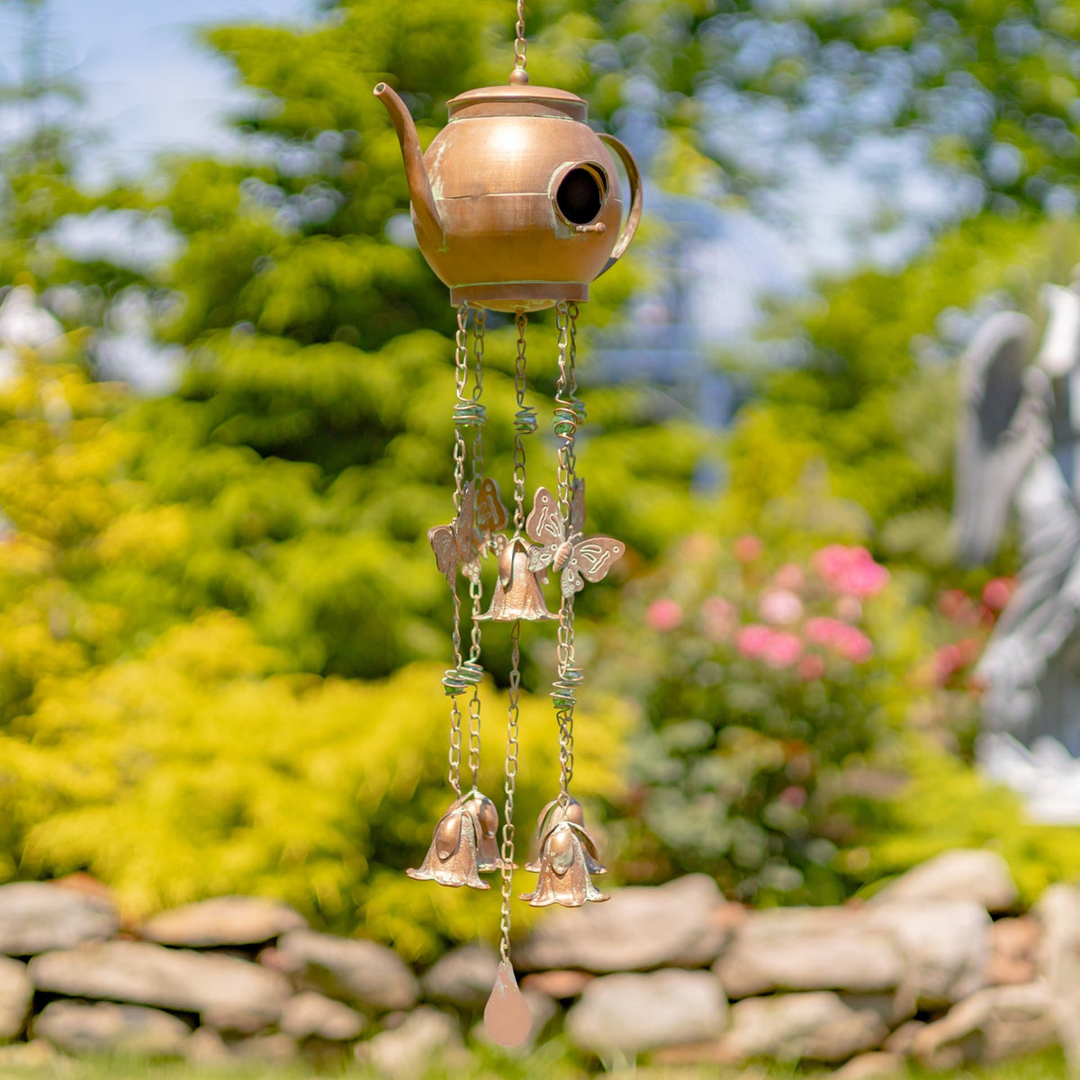 A hanging teapot wind chime with butterfly ornaments and two bells against a leafy garden backdrop.