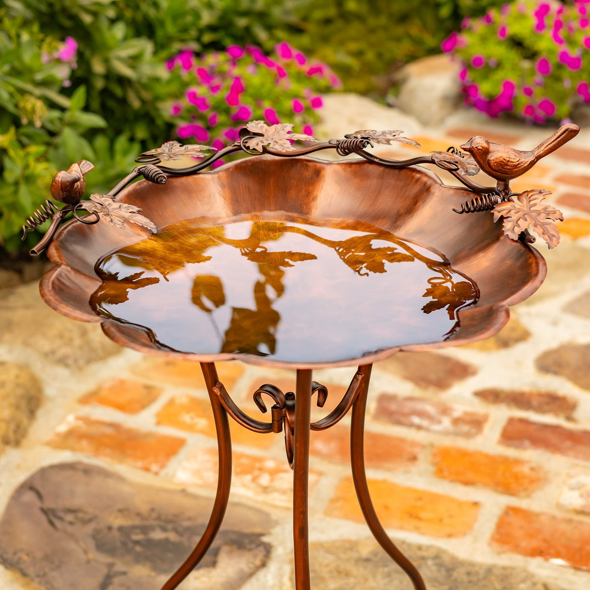 Flower-shaped bronze birdbath filled with water on a slender stand, with decorative birds and leaves around the rim.