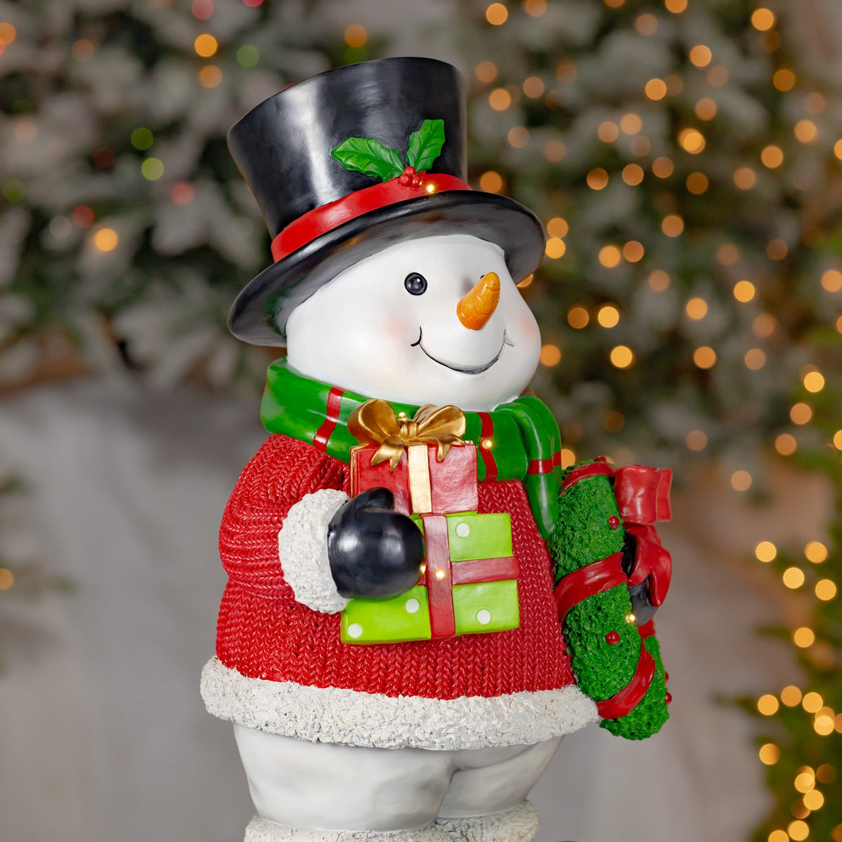 Snowman decoration viewed from the left, showing its presents and wreath against a blurred Christmas tree.