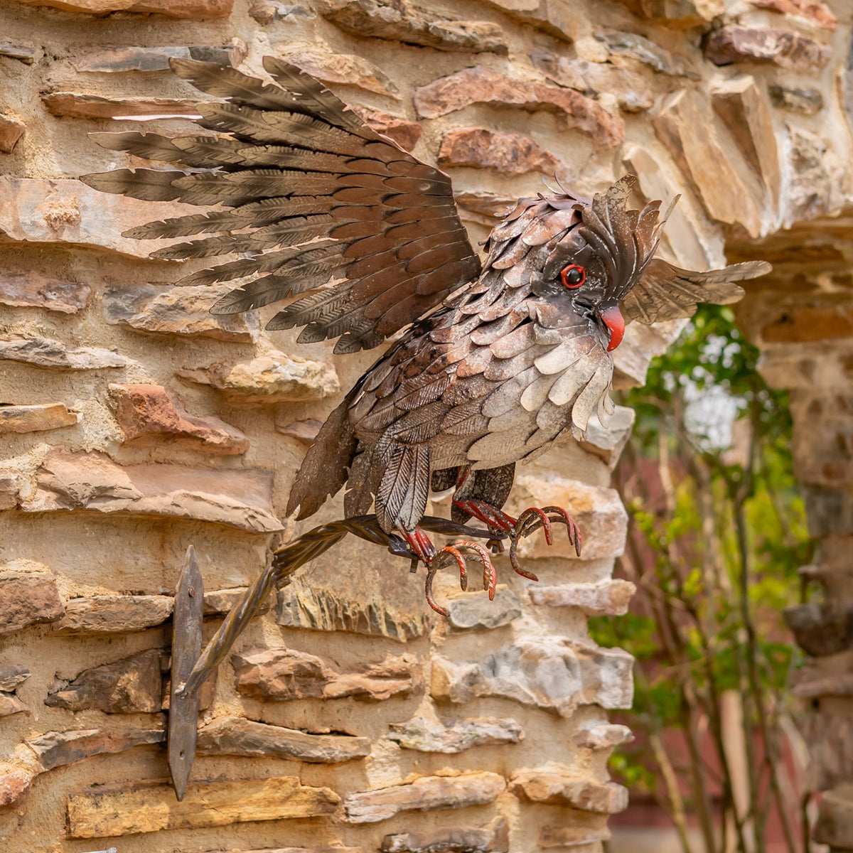 Angled view of a metal owl taking flight from a branch, with one wing raised against stone.