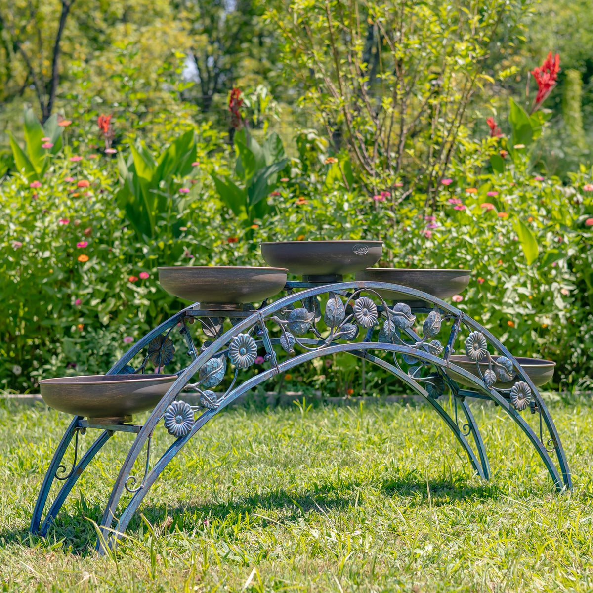 Arched metal plant stand with shallow bowls and floral scrollwork set on a lawn among garden greenery.