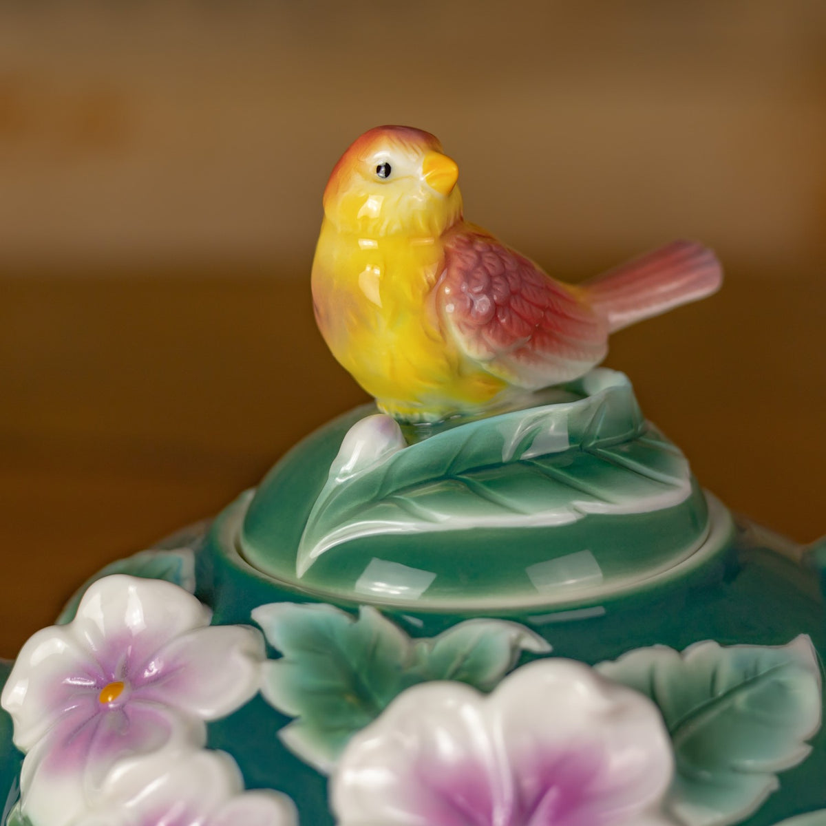 Close-up of the yellow bird perched on the leafy teal teapot lid, with flower petals visible below.