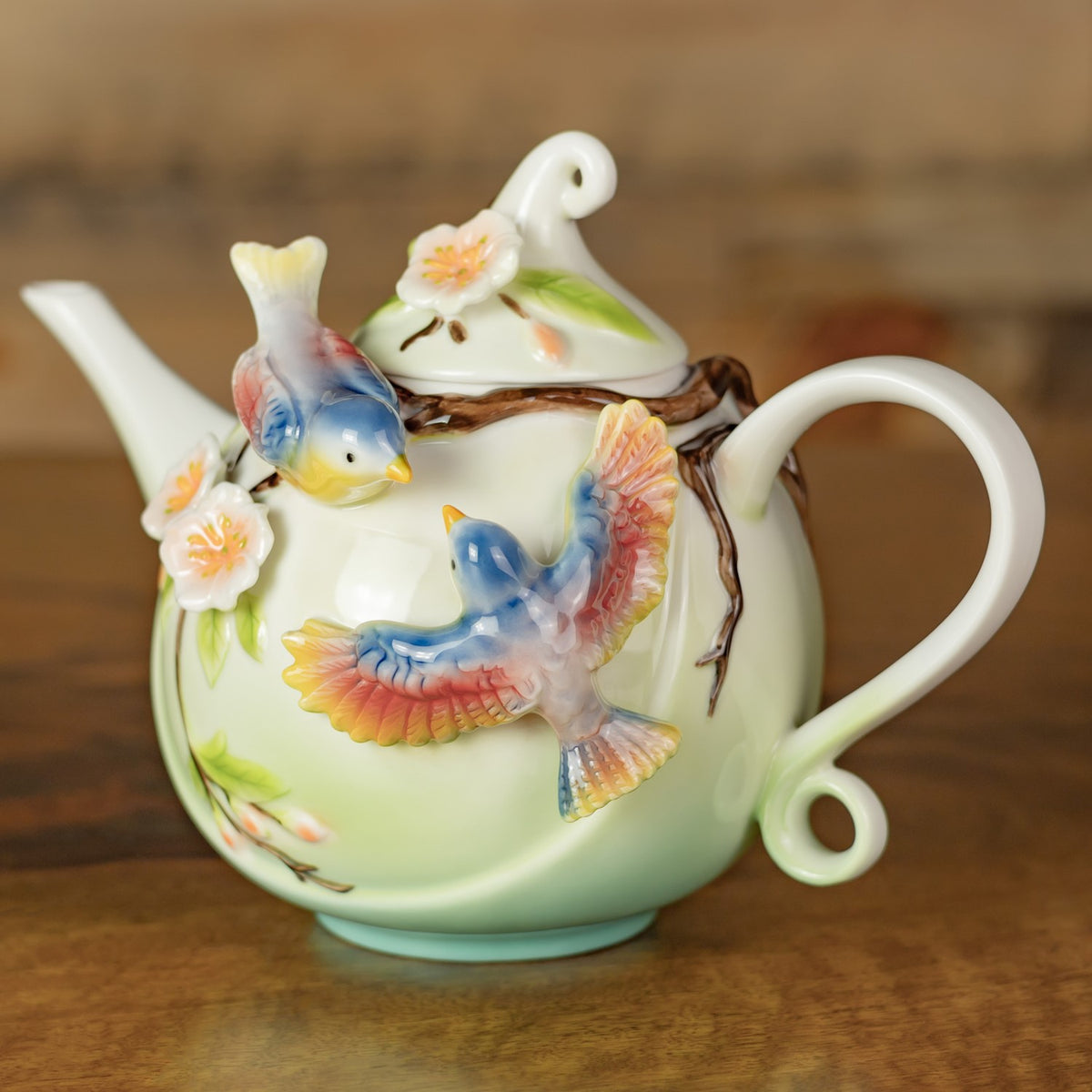Decorative teapot with two colorful birds, flowering branches, a curled lid handle, and a looped handle.