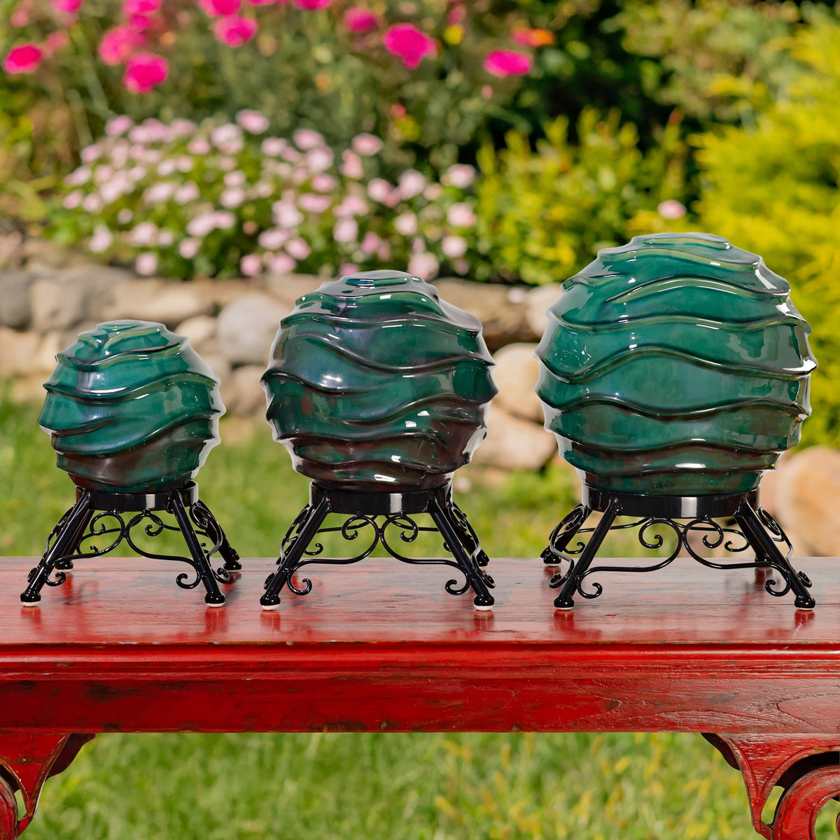 Three glossy teal globes in graduated sizes sit on ornate black metal stands atop a red outdoor table.