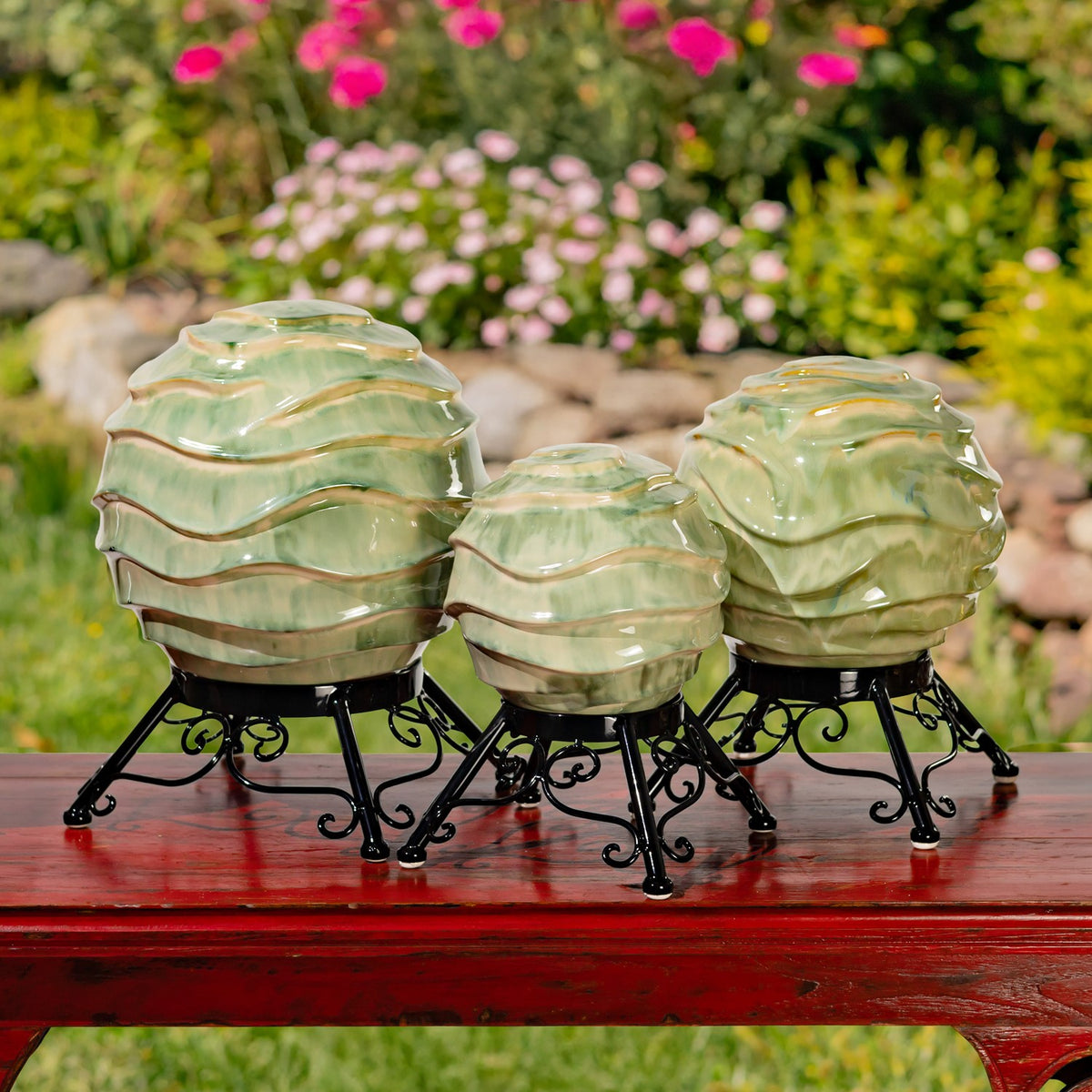 Three differently sized green glazed globes on ornate black stands displayed together outdoors.