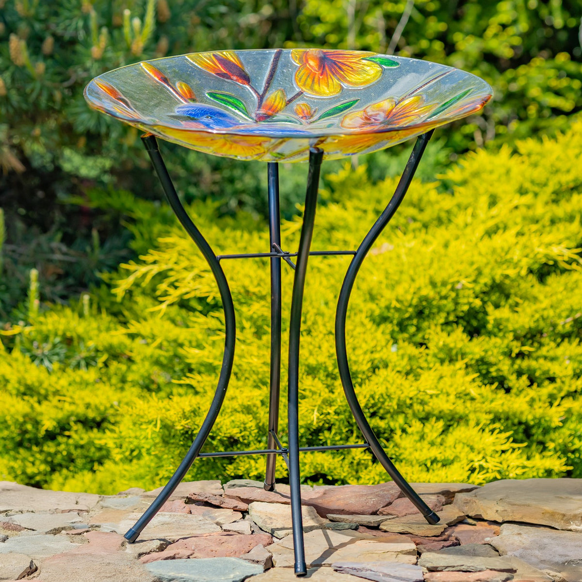 Shallow decorated glass bowl perched on a black metal stand in a garden.