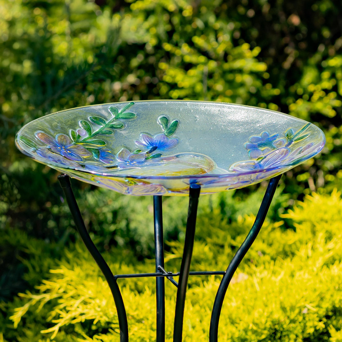 Shallow glass birdbath bowl with raised floral details resting on a dark metal stand in a garden.