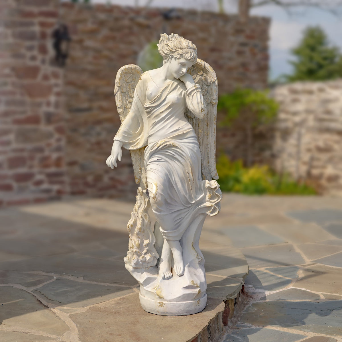 The full angel statue stands on a round base on a stone patio, with wings and draped robes.