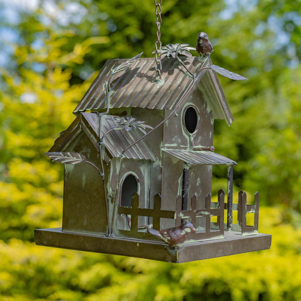 Angled view of a hanging birdhouse with corrugated roofs, leafy metal accents, arched entrances, and a fenced base.