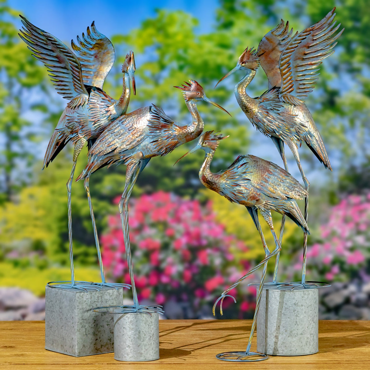 Four metal heron sculptures in varied poses, displayed on individual bases in a garden setting.