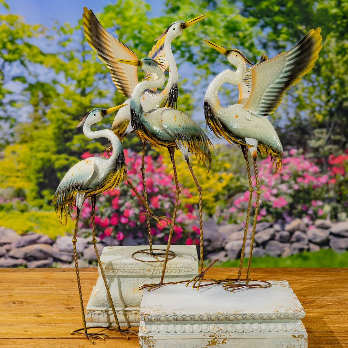 Four metal crane figurines, including two with raised wings, stand together on a wooden surface outdoors.