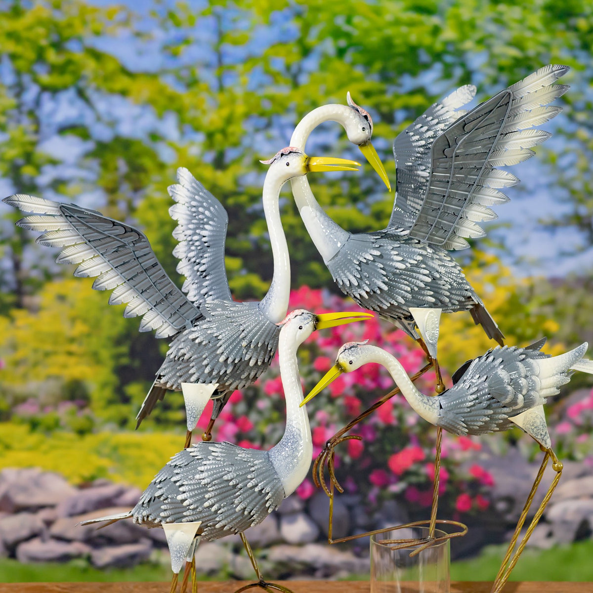 Four metal heron figures arranged together outdoors, with varied poses and wings spread against greenery.