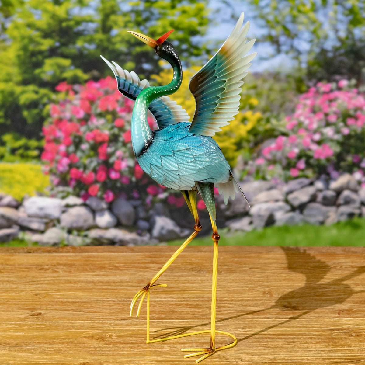 A blue heron figurine stands on long yellow legs with one wing lifted against a garden backdrop.