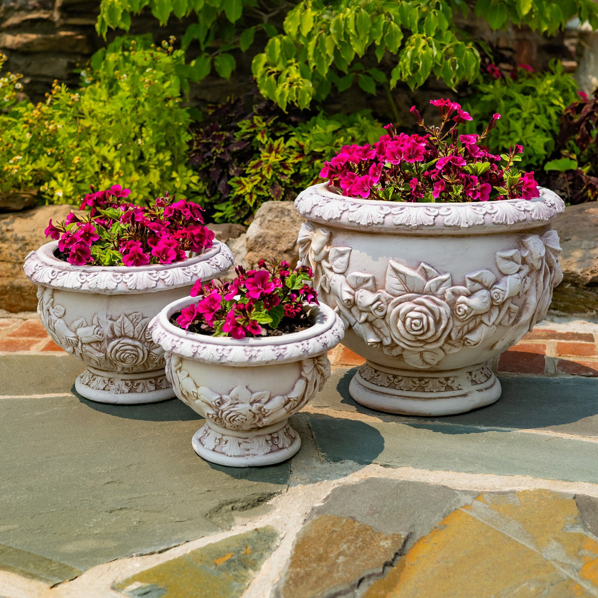 Three floral-relief urn planters in different sizes, arranged on a stone patio with bright pink blooms.
