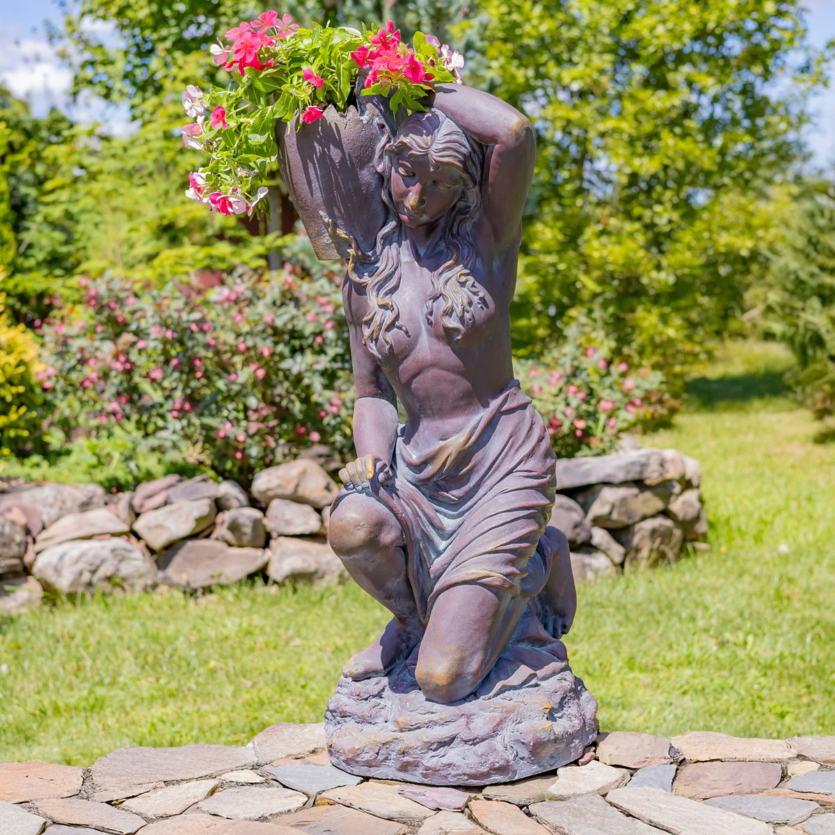 Kneeling statue with pink flowers spilling from the urn, pictured beside a low stone border.