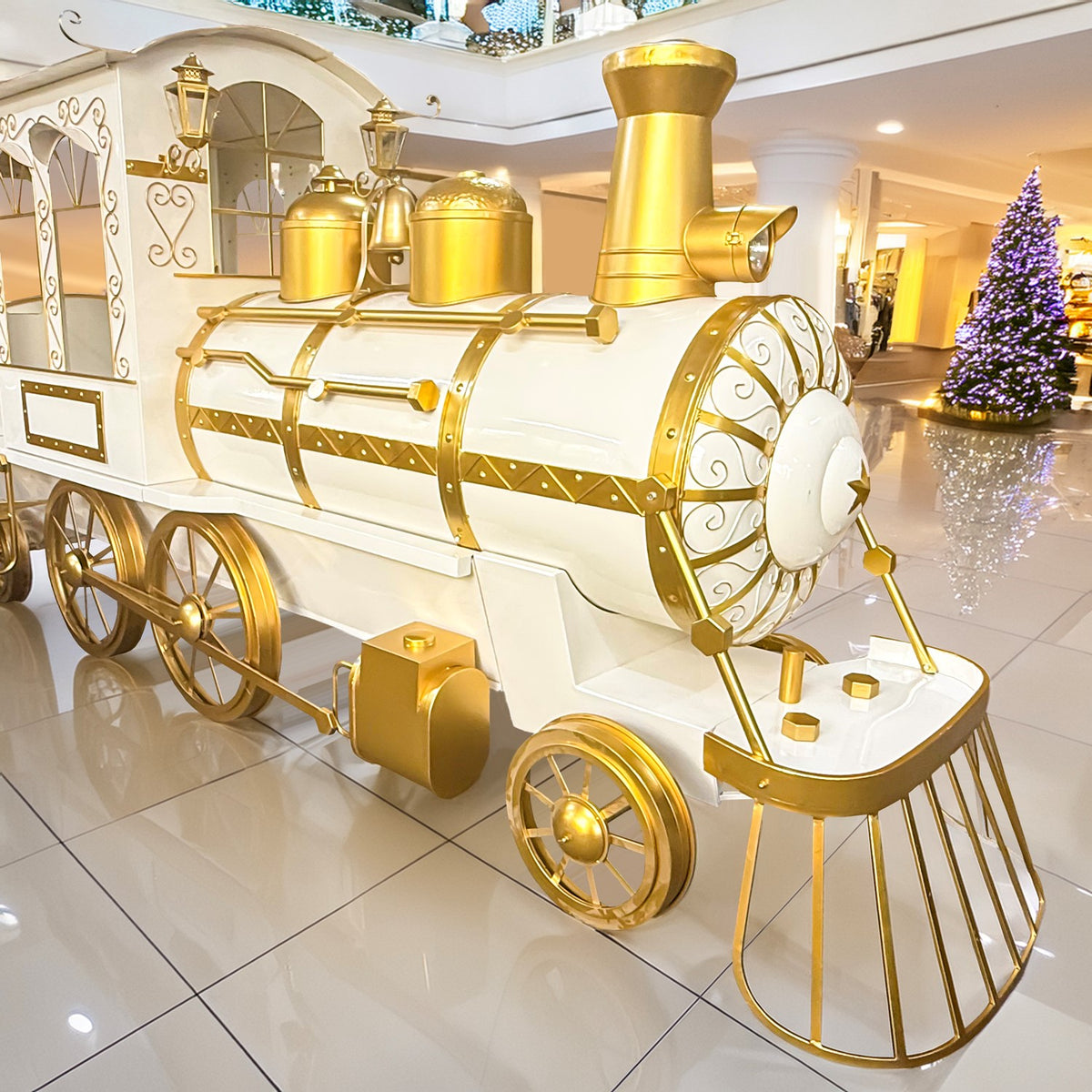 White and gold decorative train displayed in a bright indoor hall, with a rounded front grille and large wheels.