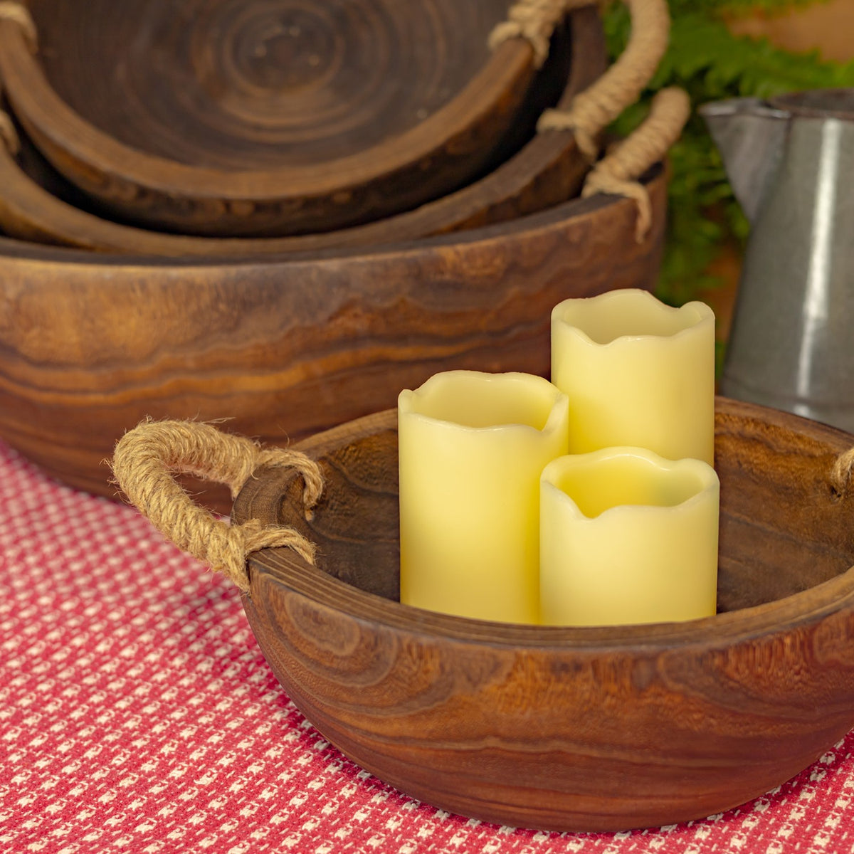 A wooden tray with a thick rope handle holds three yellow candles, with nested trays behind it.