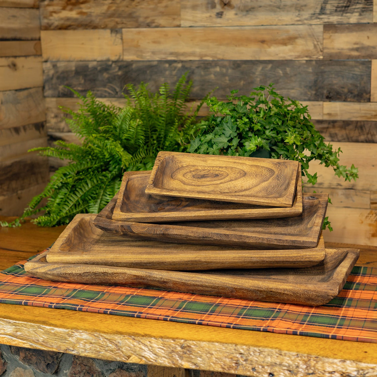 Five empty wooden trays stacked by size on a plaid cloth against a wood-paneled wall.