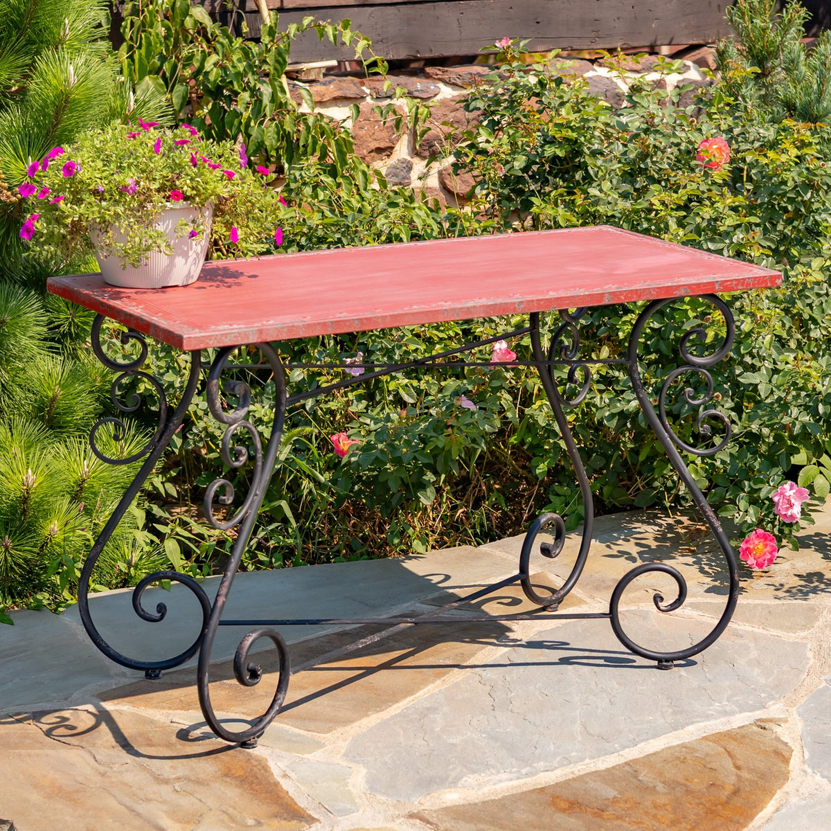Red rectangular table with a dark, ornate scrollwork frame on a stone patio among flowering plants.
