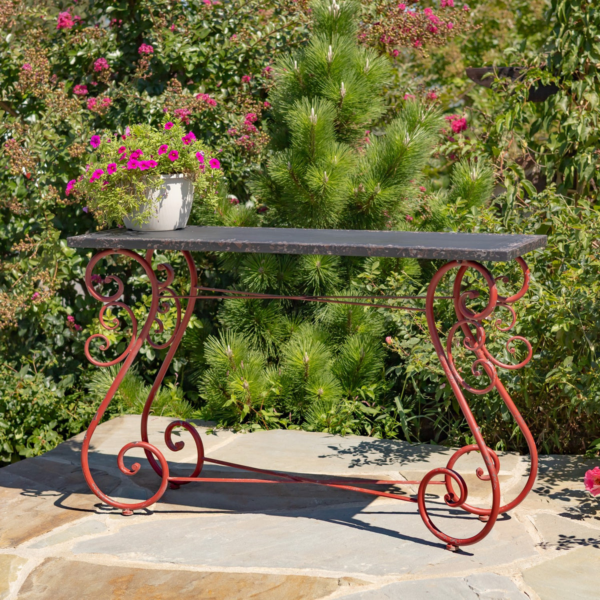 Side view of a dark rectangular table with red scrollwork legs, set on a stone patio among greenery.