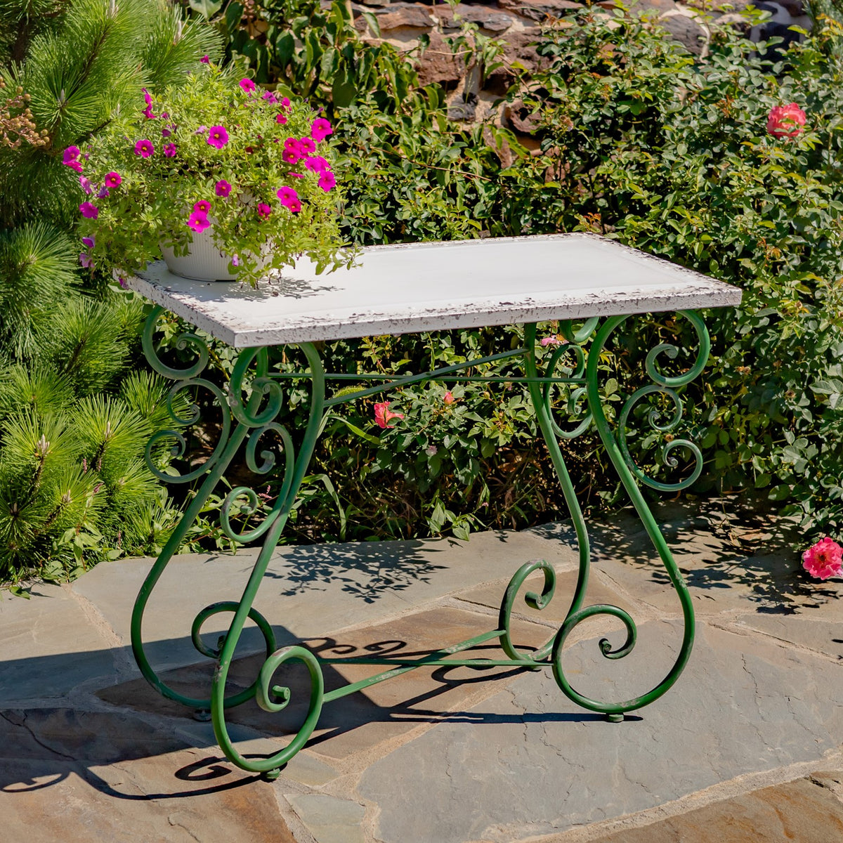 White rectangular tabletop and curled green iron legs viewed from the side on a stone patio.