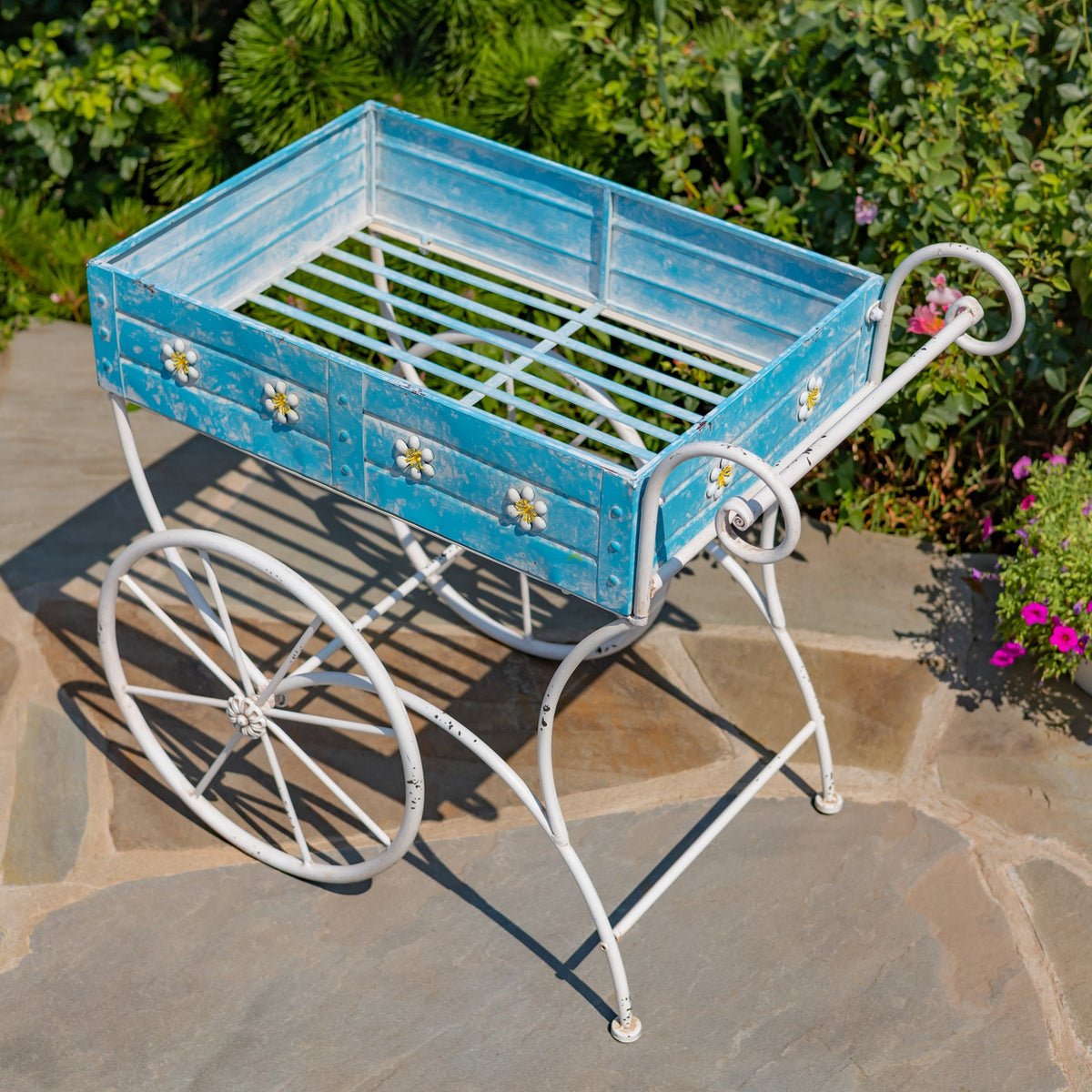 An empty blue flower cart viewed from above, showing its slatted base, white frame, wheels, and curled handle.
