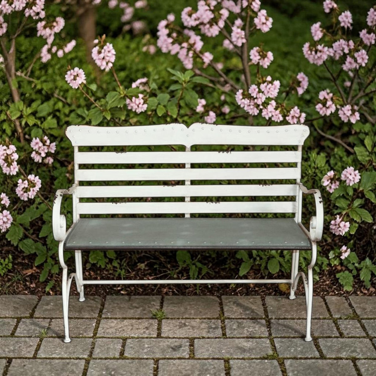 White garden bench with horizontal back slats sits on a paved path beneath flowering branches.