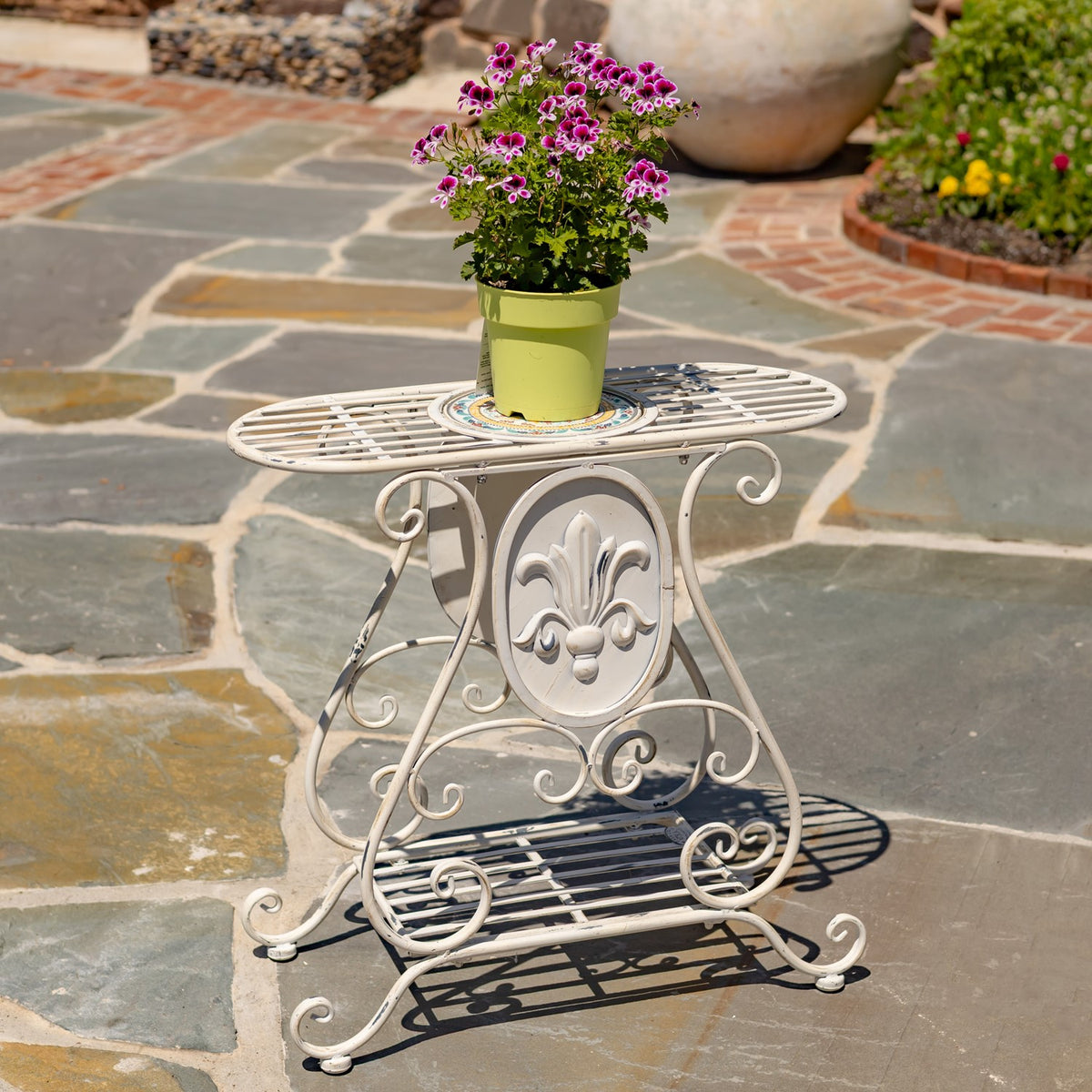 A cream scrollwork plant stand with a potted flowering plant on its oval slatted top.