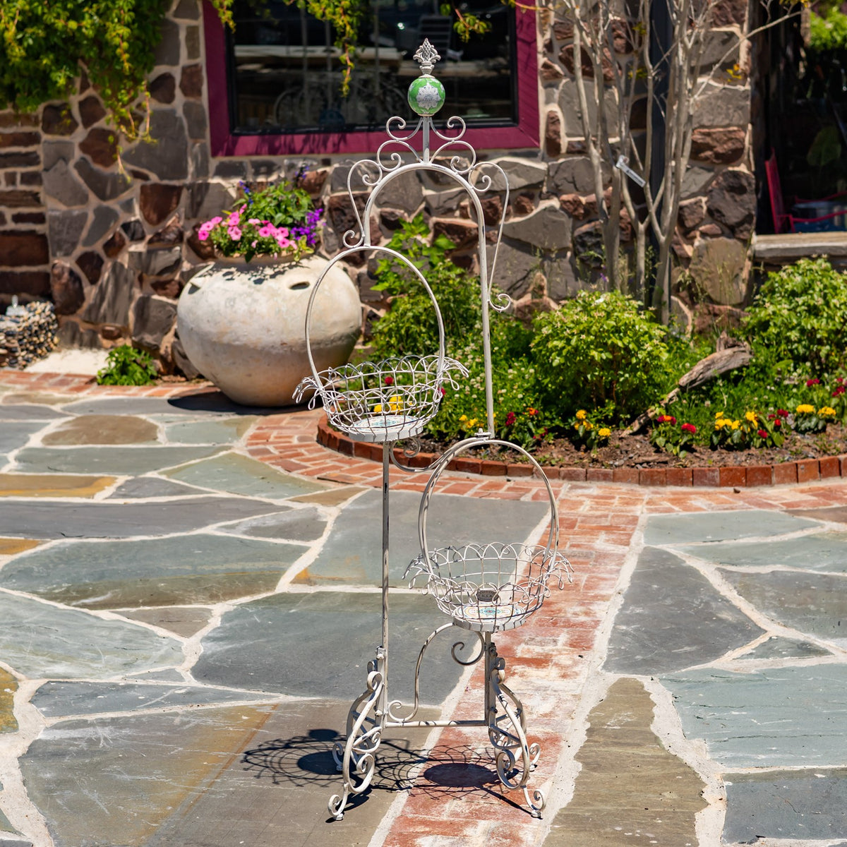 Two empty wire baskets hang at different heights on the decorative stand, shown on a garden patio.