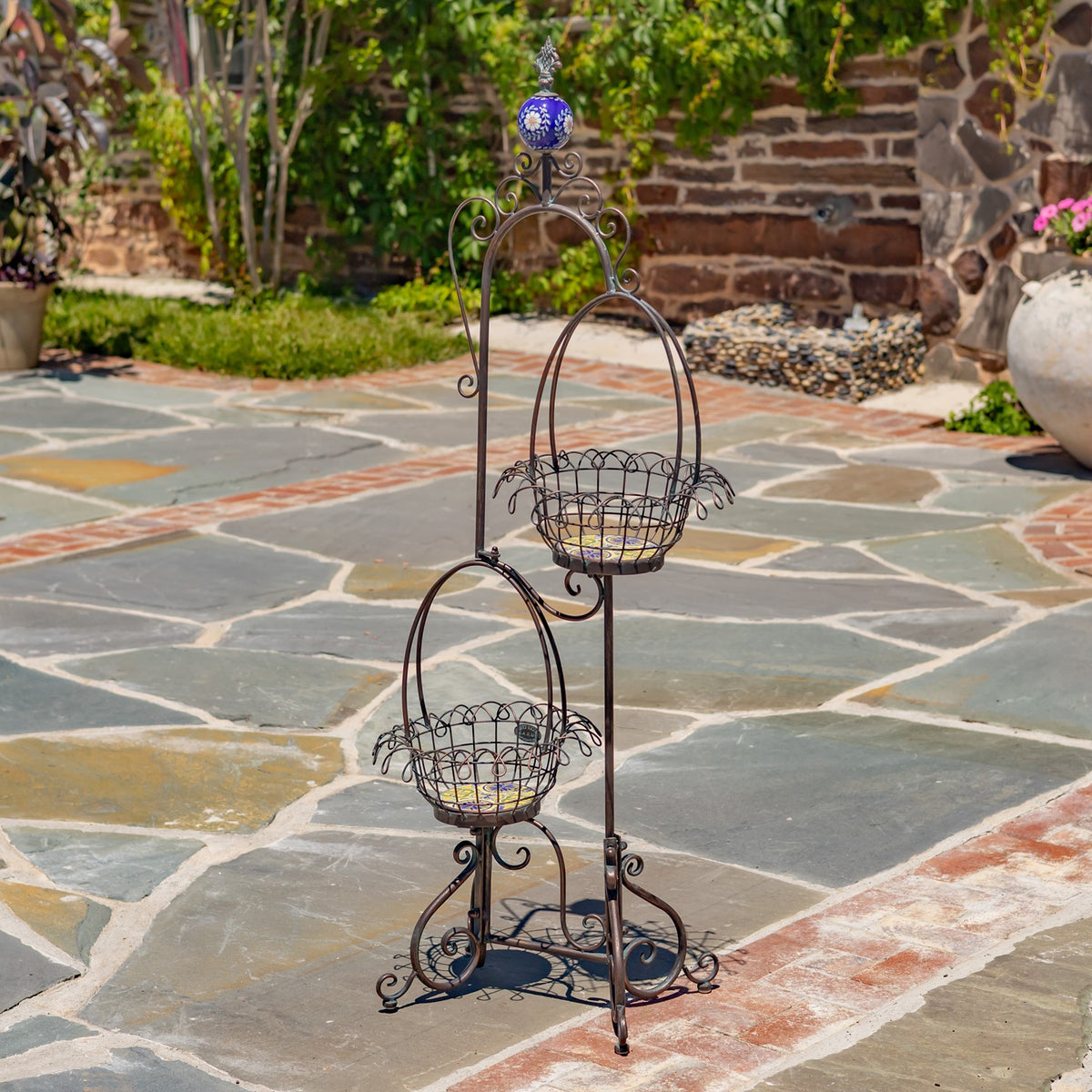 Empty two-tier wire basket plant stand with curling metalwork and a blue finial on a stone patio.