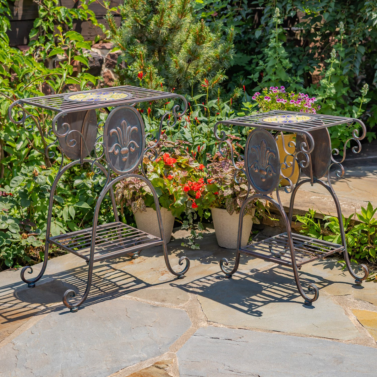 Pair of decorative iron plant stands shown from the side among garden greenery and potted flowers.