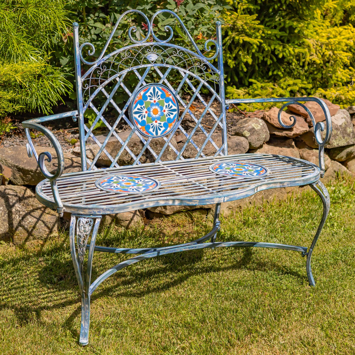 Three-quarter view of a metal garden bench with scrollwork arms and floral mosaic details.
