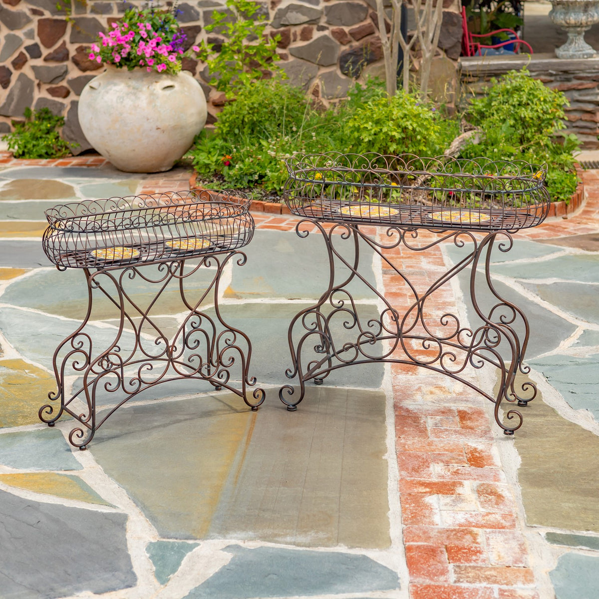 The two empty oval plant stands show their mosaic tops and curled metal legs on a stone patio.