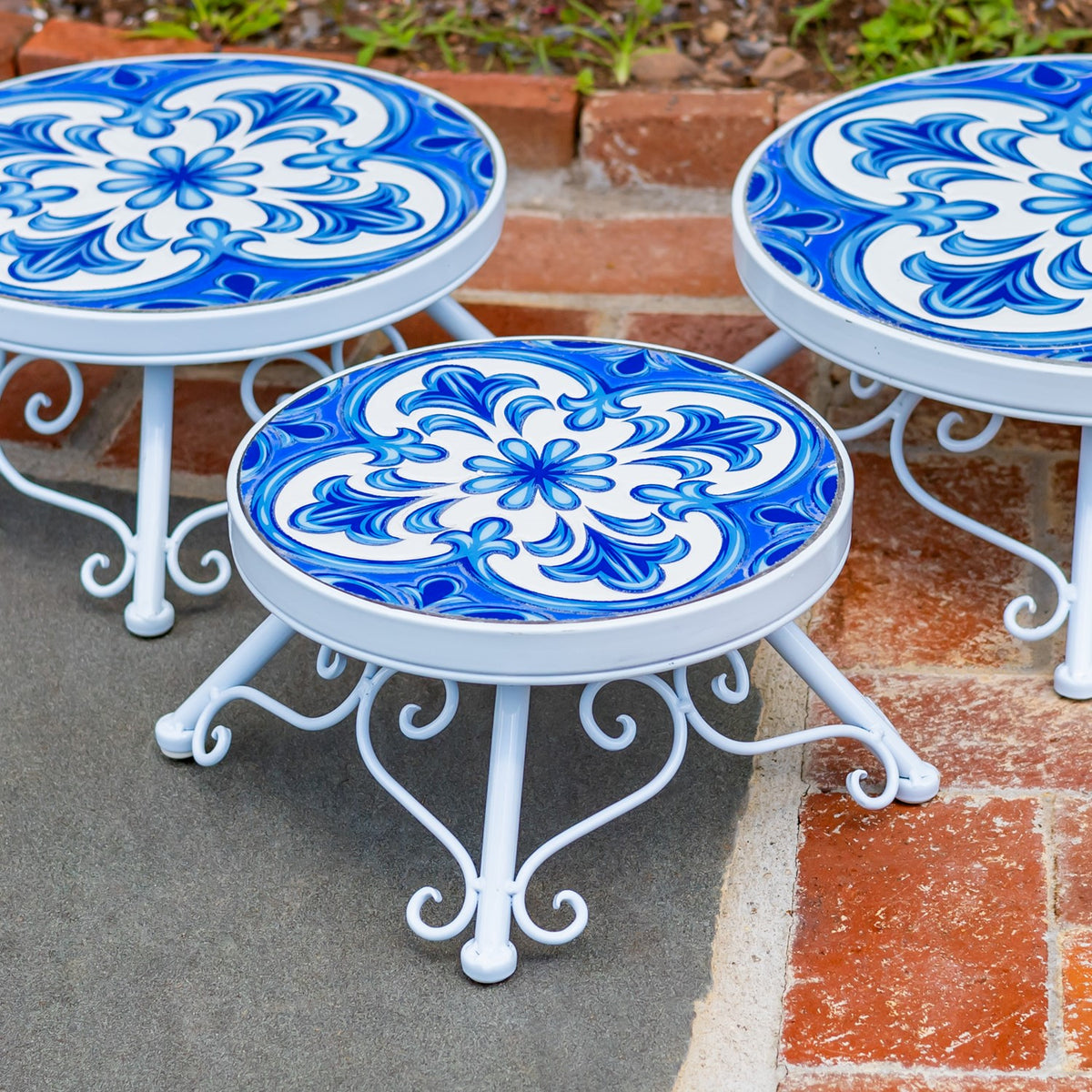 Three round stands with ornate blue-and-white tops and curled white metal legs on a patio.