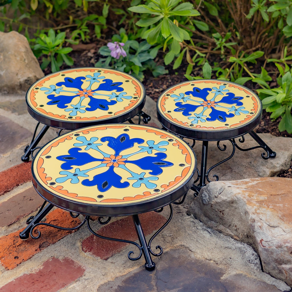 Three round mosaic plant stands in different sizes sit together among garden rocks and greenery.
