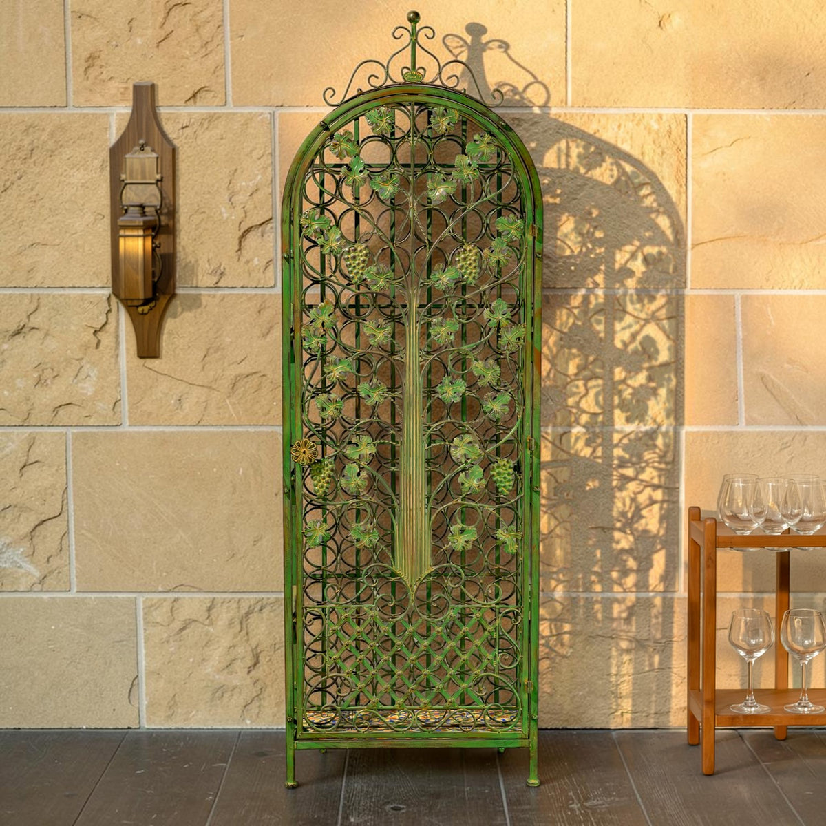 Front view of an ornate arched iron wine rack against a stone wall, with a small glassware stand beside it.