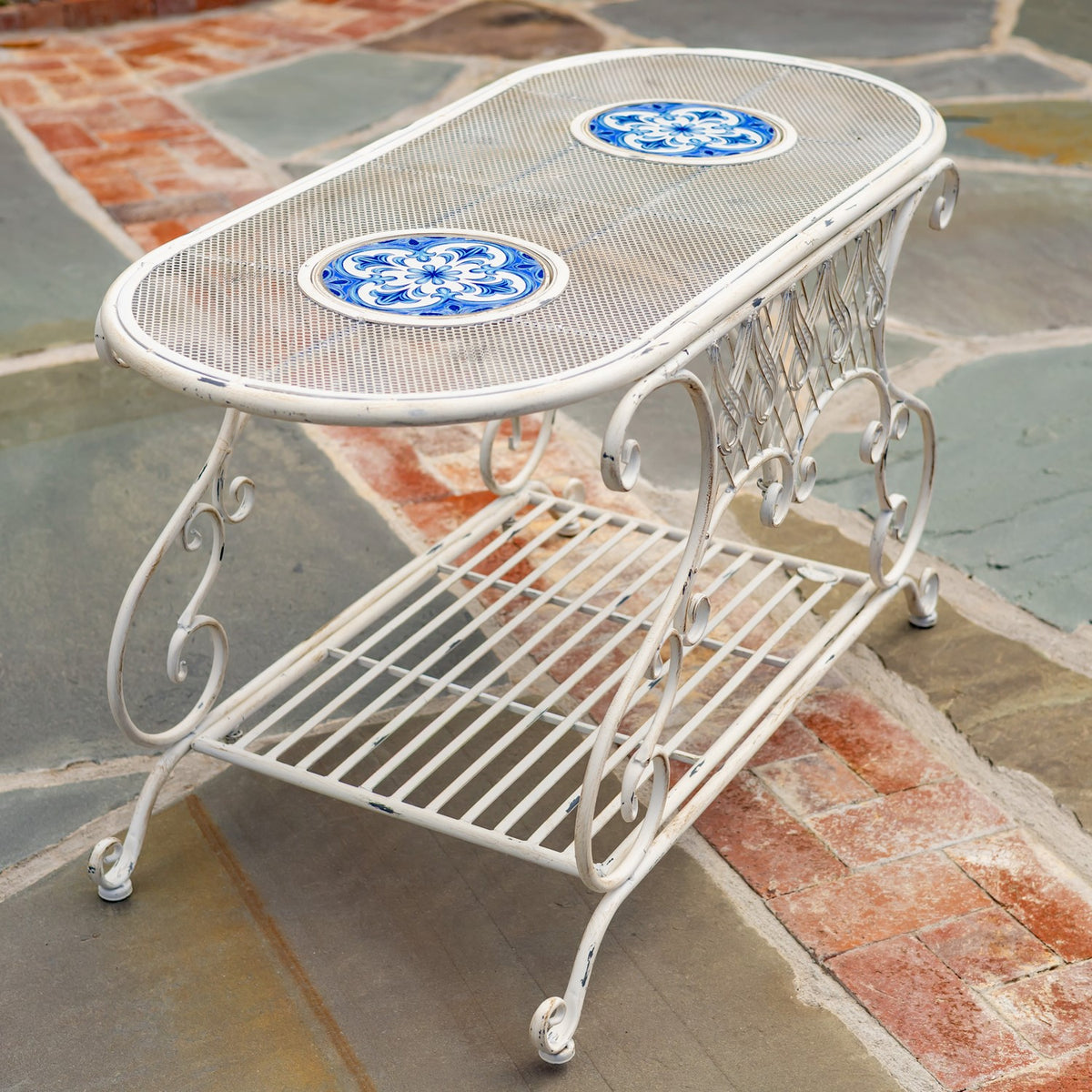 Angled view of the table showing its mesh oval top, two blue mosaic insets, curled legs, and slatted shelf.