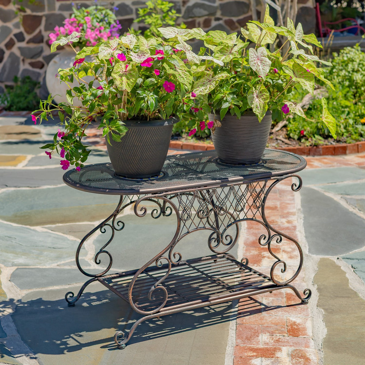 Oval metal garden table with mosaic accents and lower shelf, topped with two potted flowering plants.