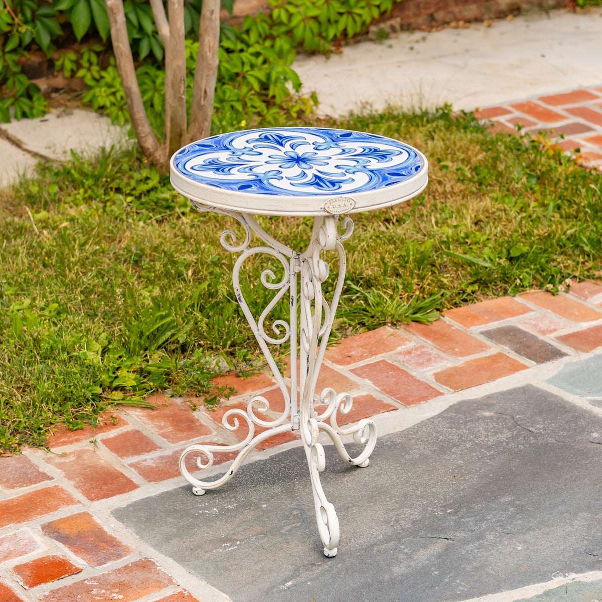Empty white metal plant stand with a round blue-and-white patterned top on a brick garden path.