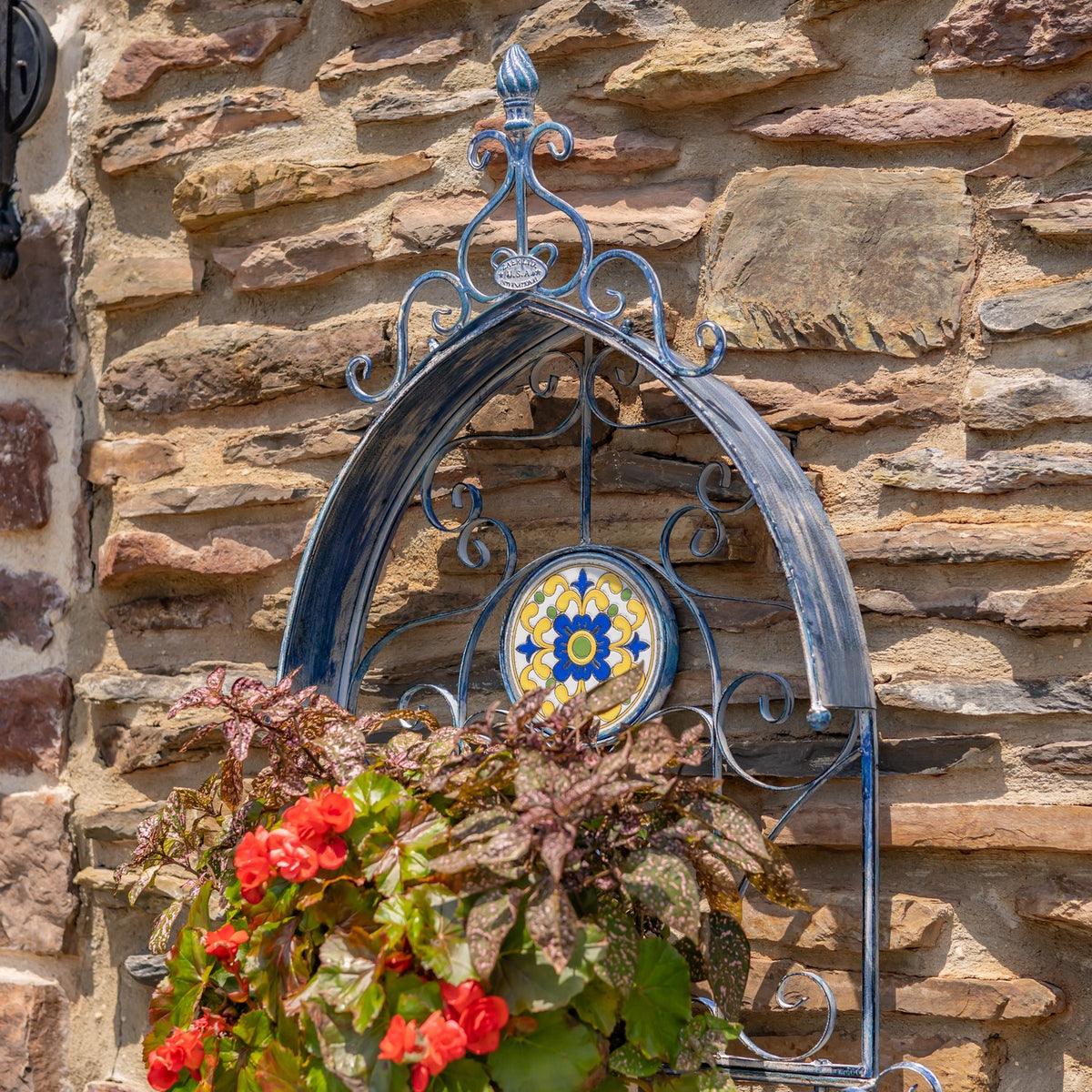 Flower-filled iron wall planter with a blue, yellow and white mosaic medallion on a stone wall.