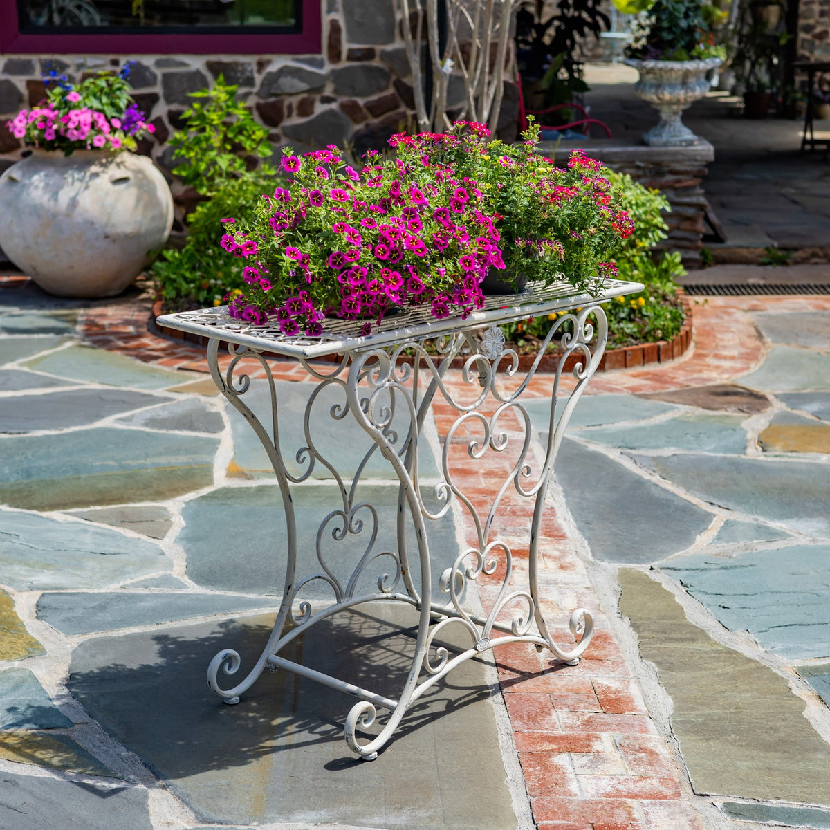 Three-quarter view of the ornate metal table on a stone patio, with pink flowers arranged on top.