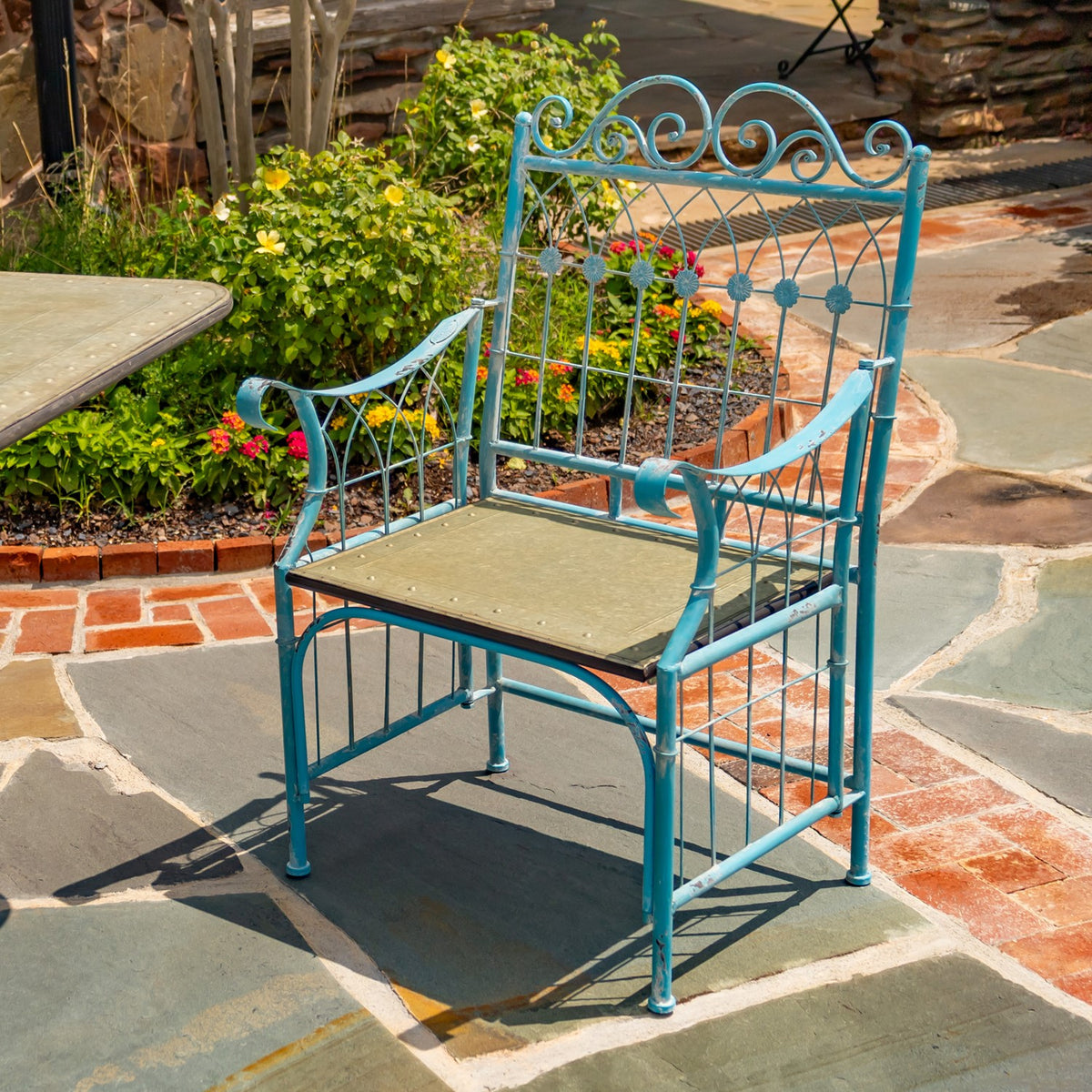 Turquoise metal dining chair with a tan seat on a stone patio beside a garden and partial table.