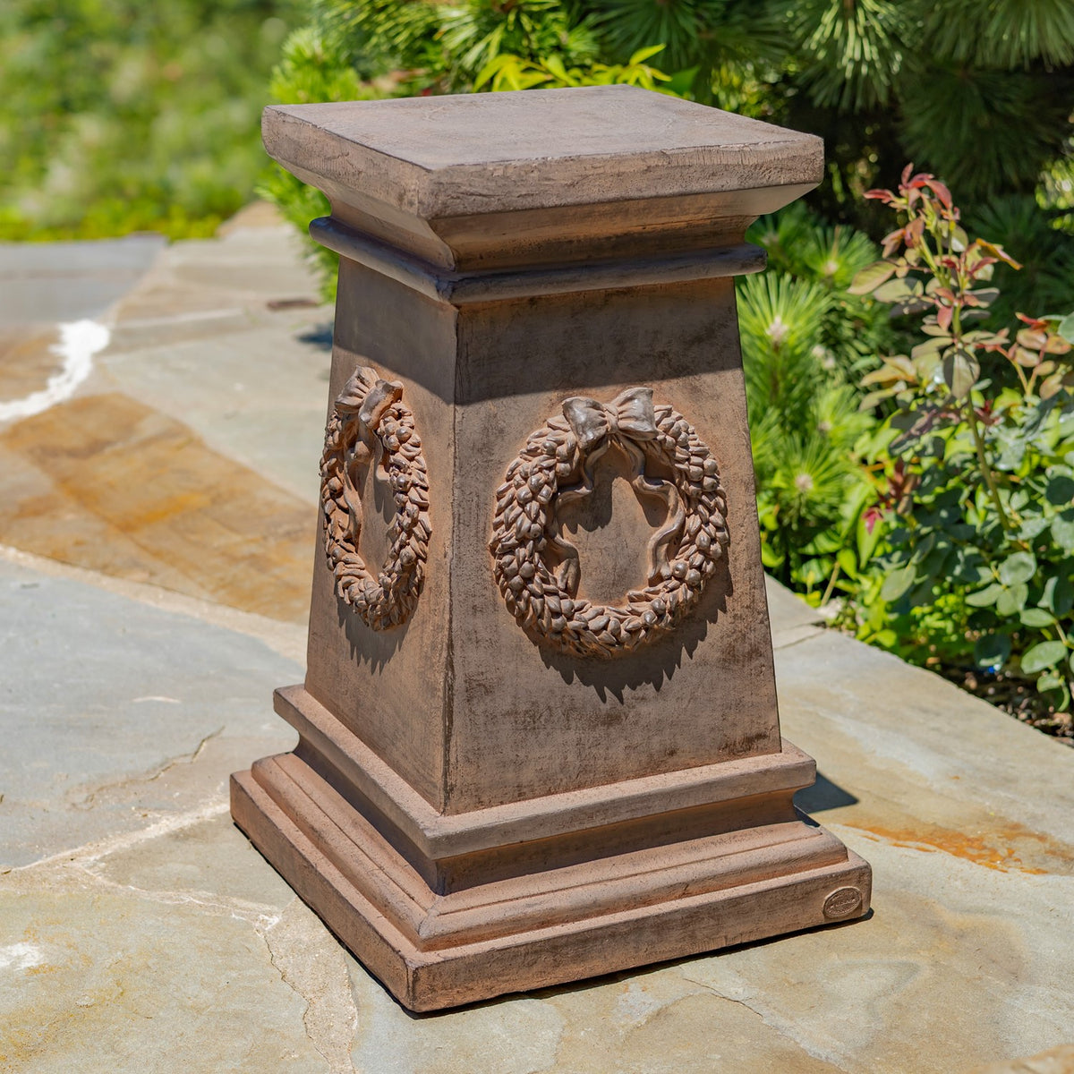 Brown square pedestal with a broad top and raised wreath decorations, shown outdoors on a stone path.