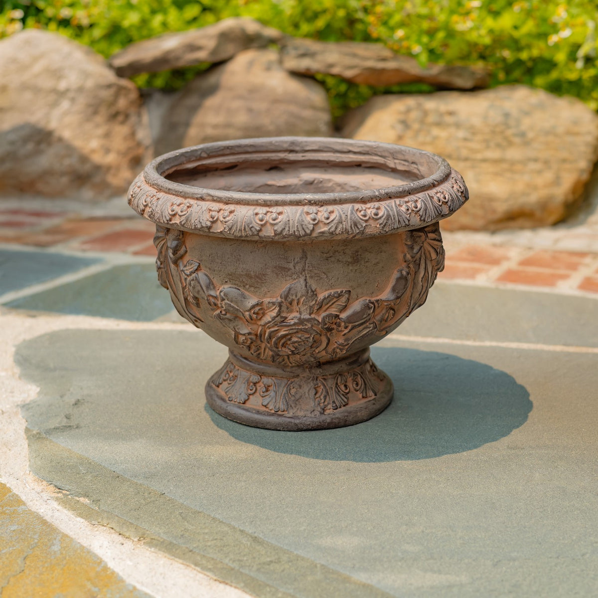 An empty round planter with a raised floral design and decorated rim on a stone patio.