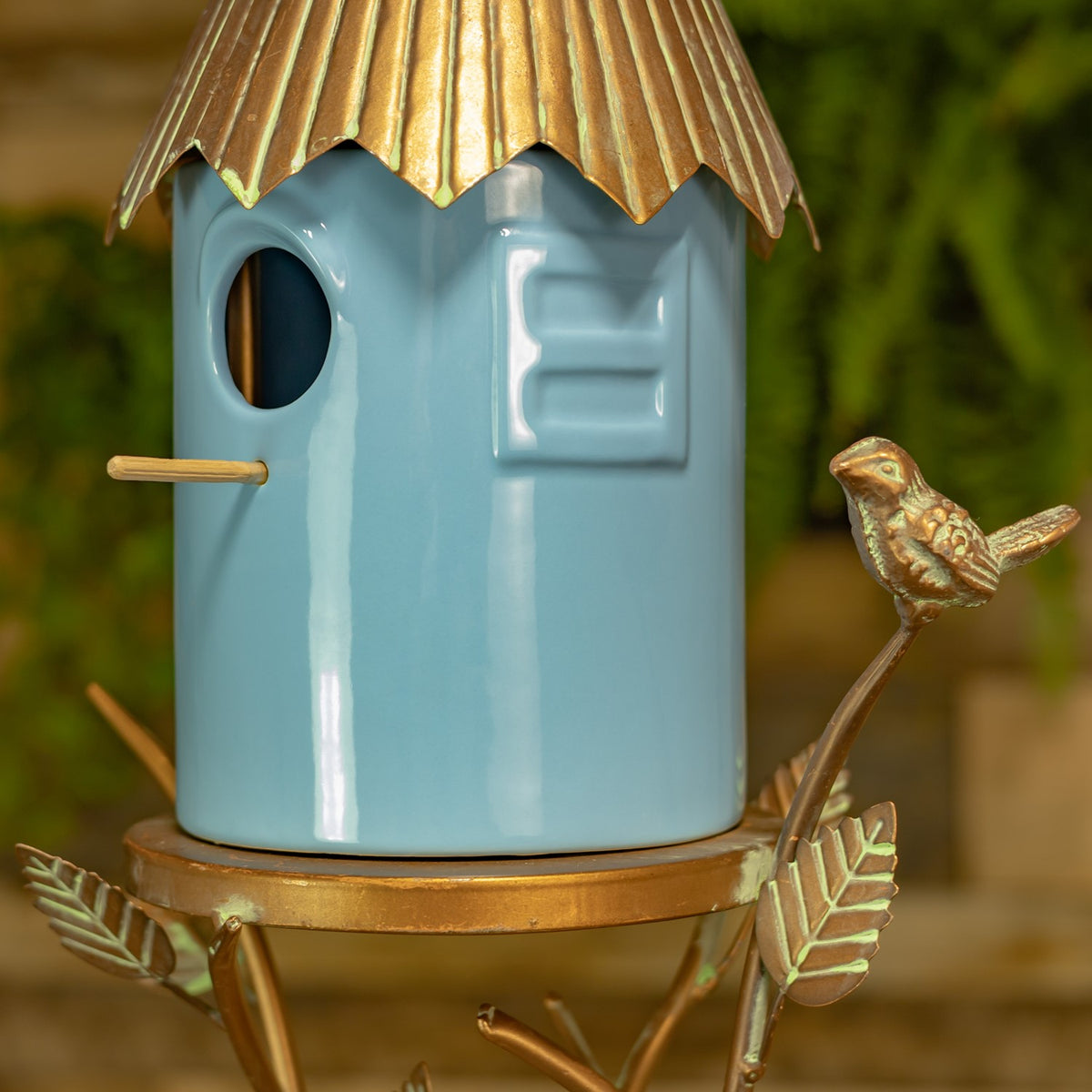 Close-up of the blue birdhouse entrance, gold-toned roof edge, perch, and decorative bird on the frame.