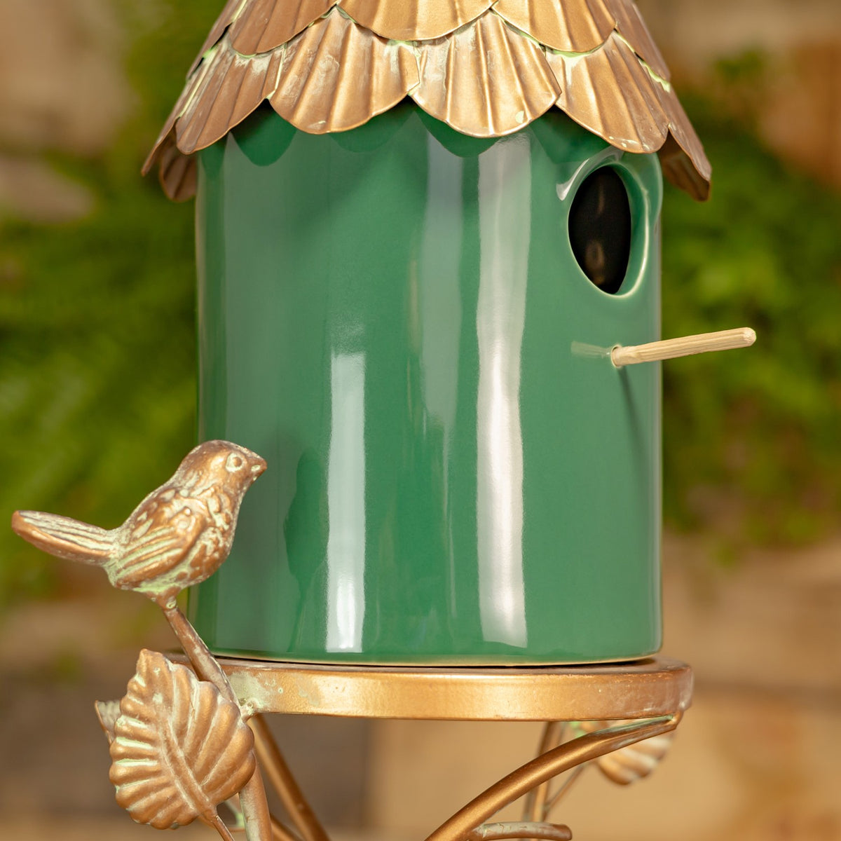 Close-up of the green birdhouse body, round entrance hole, short perch, and bird ornament beneath the copper-tone roof.