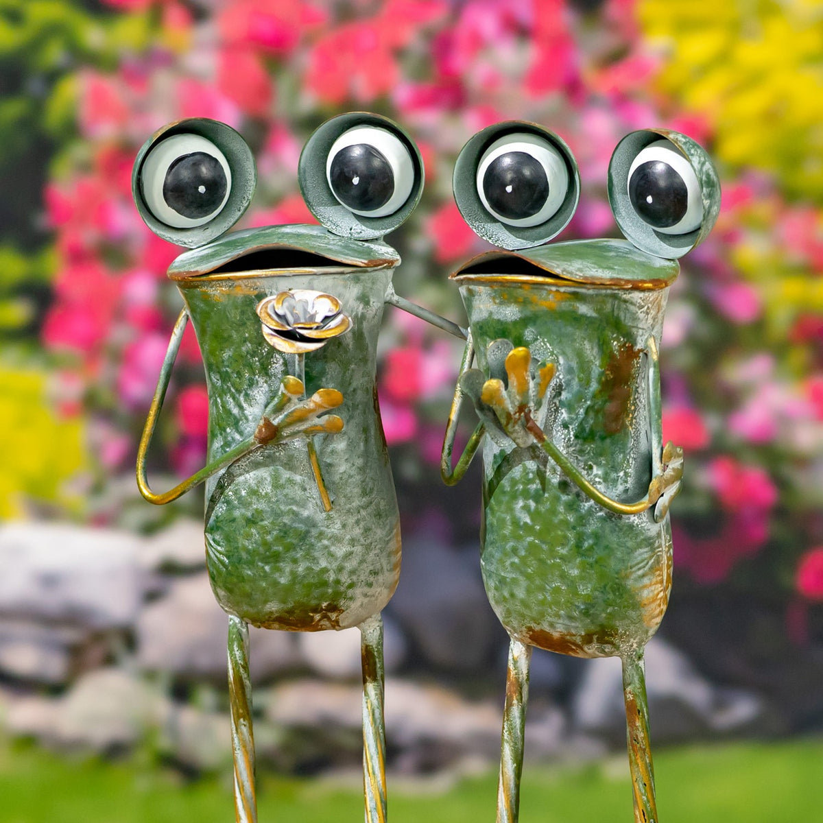 Two green metal frogs with oversized eyes and long wire limbs stand side by side against a garden backdrop.