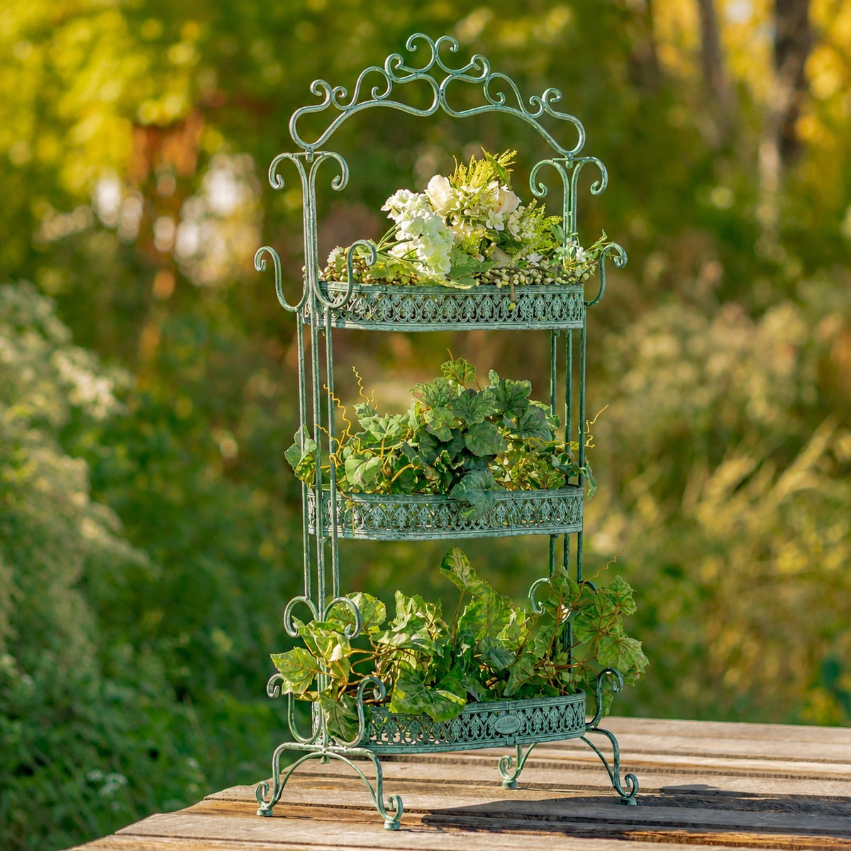 Green iron plant stand displaying flowers and leafy plants across its three tiers outdoors.